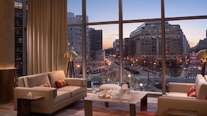 A modern hotel room with a large city view, a sofa, a coffee table, and a lamp.