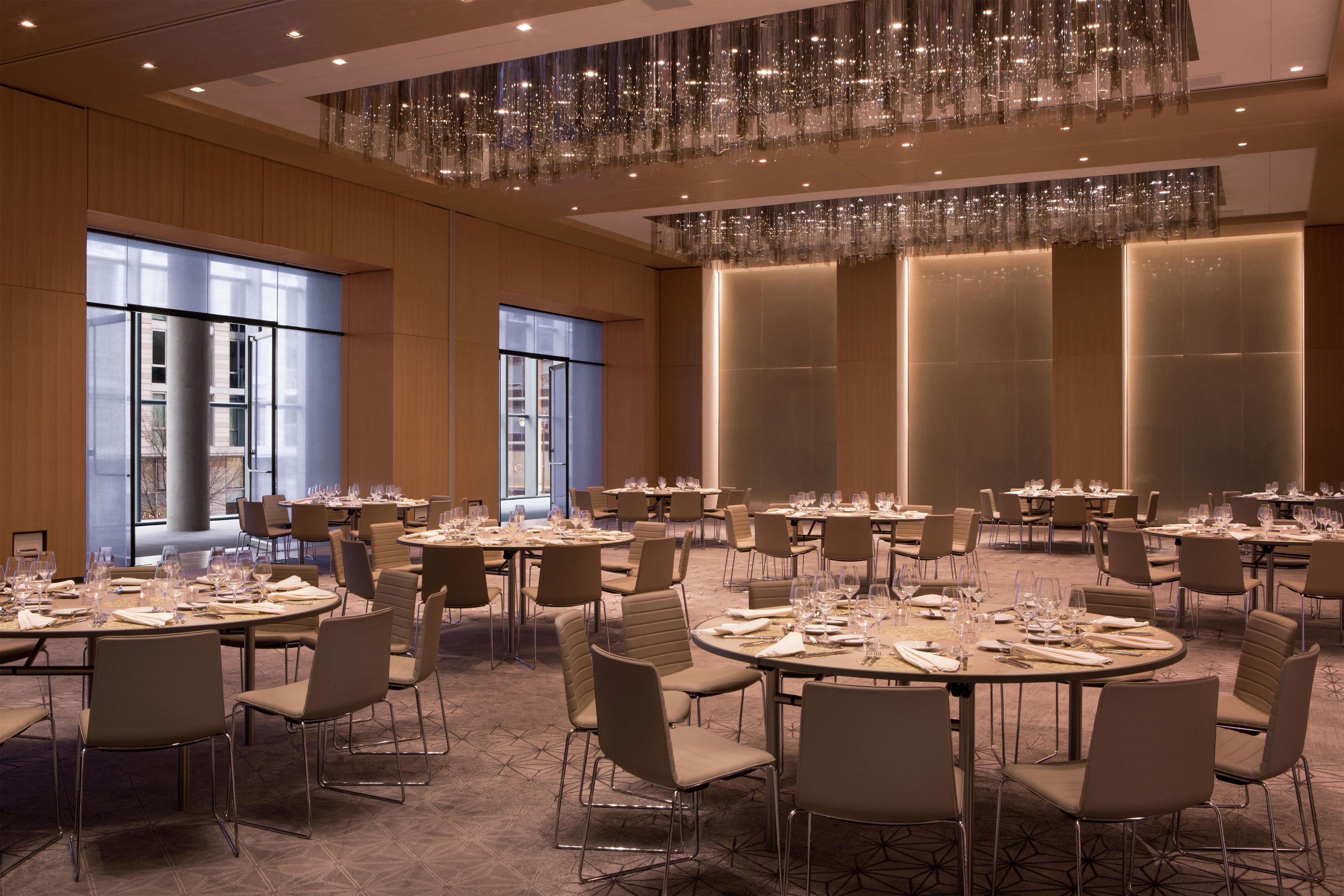 A spacious banquet hall with round tables set for dining, large windows, and a modern chandelier.