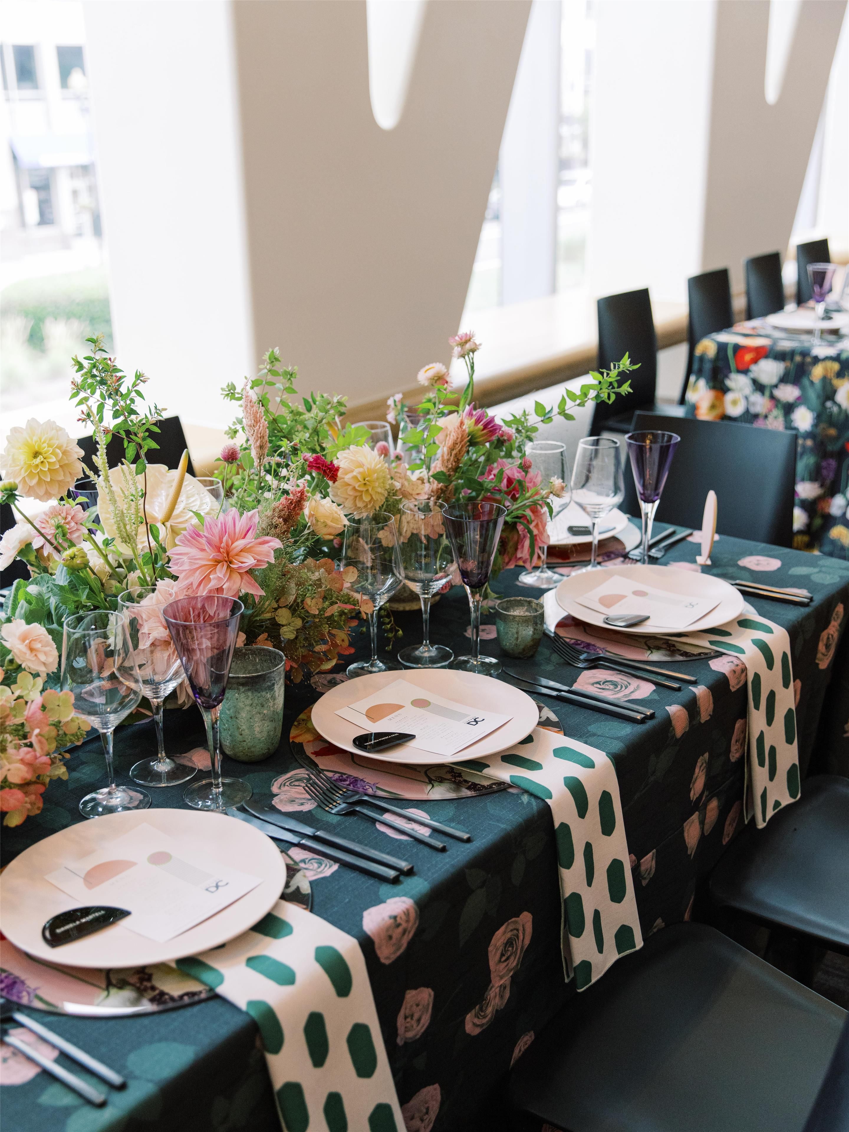 Una mesa dispuesta para una comida, con centros de mesa florales, copas de cristal y un mantel estampado con lunares.