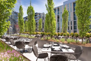 Un área de comedor al aire libre con mesas y sillas, rodeada de árboles y edificios modernos.