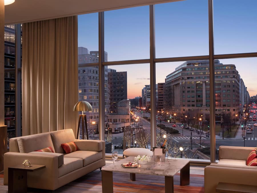 A modern hotel room with a large city view, a sofa, a table with drinks, and a lamp.