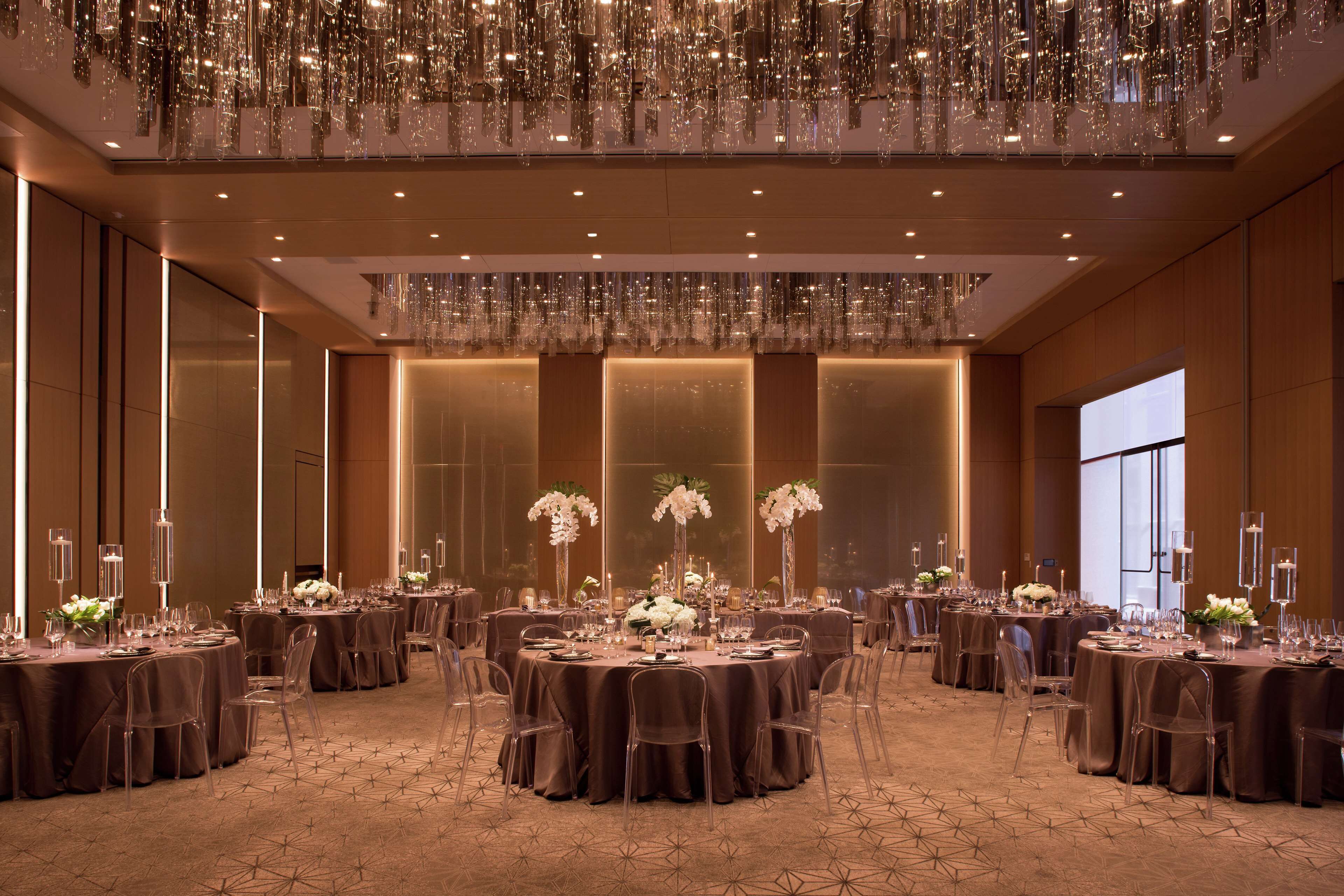 A banquet hall with round tables, clear chairs, and a chandelier.