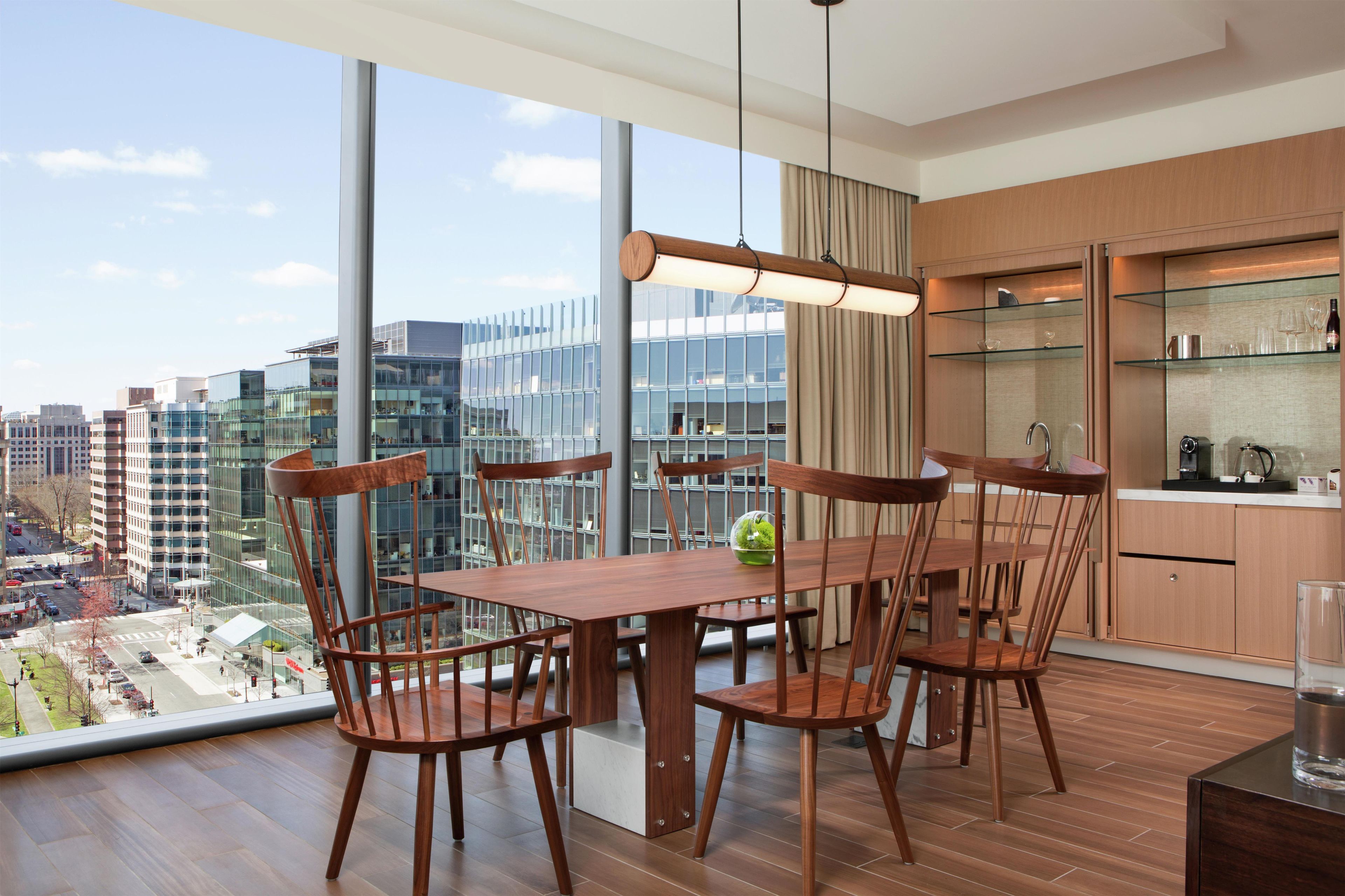 Un comedor moderno con mesa y sillas de madera, ventanas amplias con vista a la ciudad y una cocina con estantes de vidrio.