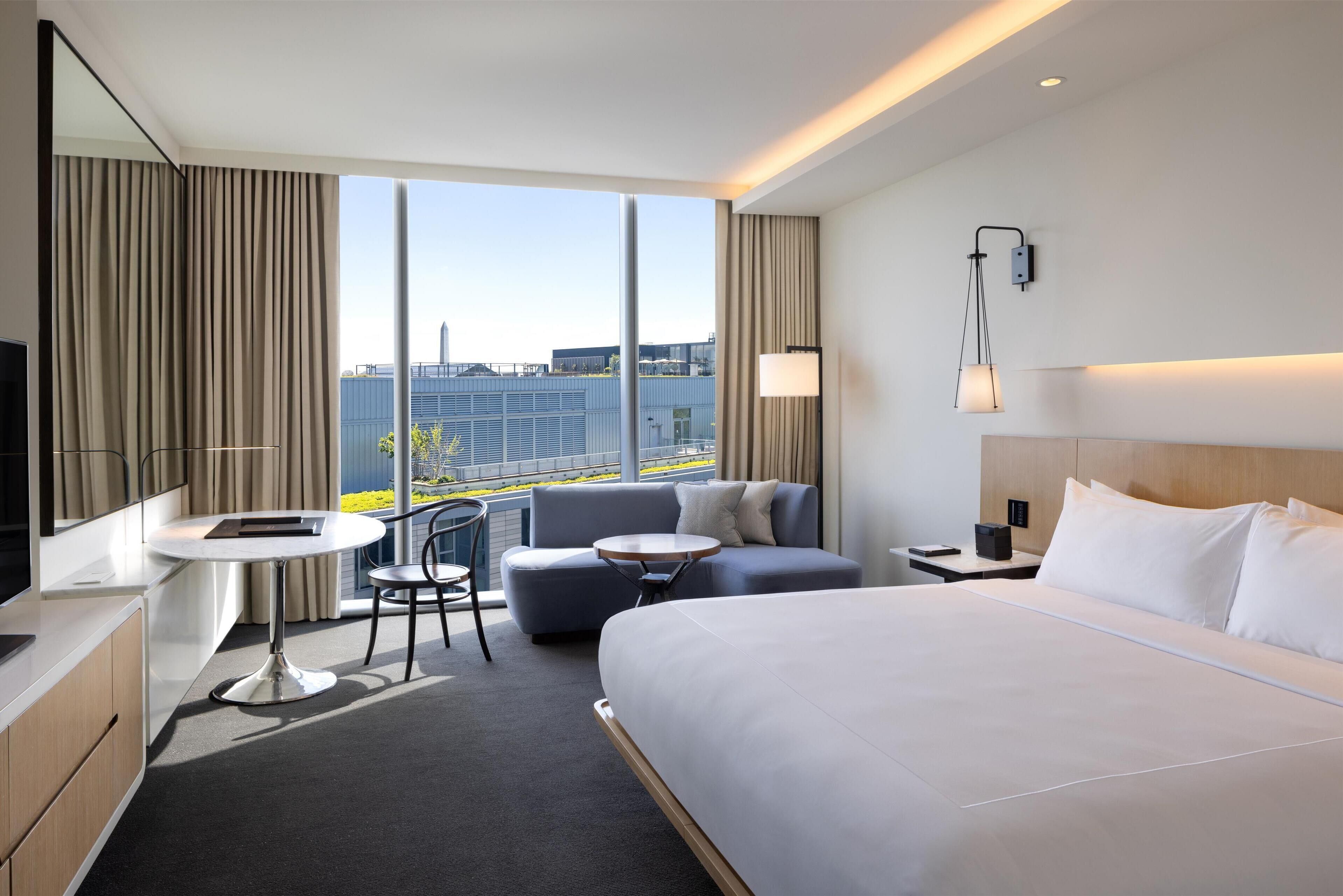 Une chambre d’hôtel moderne dotée d’un grand lit, d’un canapé, d’une petite table, d’une chaise, d’un bureau et d’une grande fenêtre donnant sur la ville.