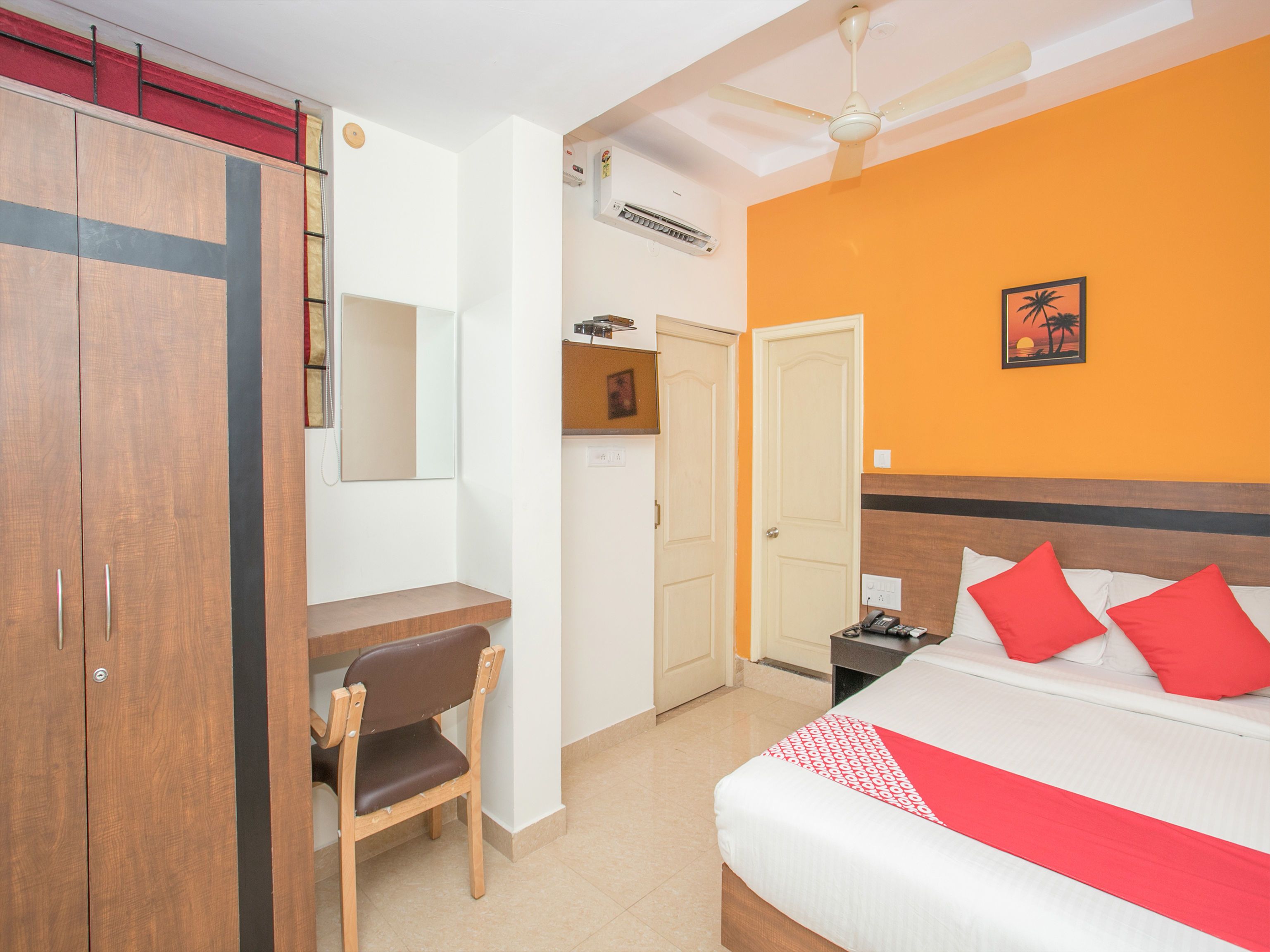 A hotel room with a bed, a desk, a chair, a wardrobe, a ceiling fan, and a wall-mounted air conditioner.