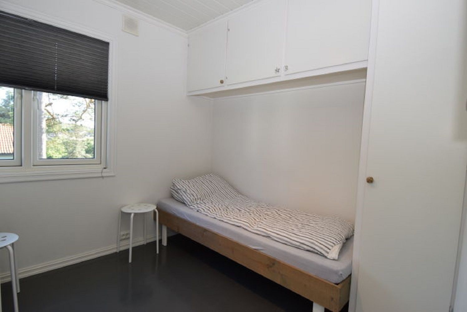 A small, single-bed room with a wooden bed frame, white walls, a window with blinds, and a small white table.