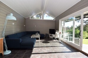 A sunlit room with a dark blue sectional sofa, a small table, a TV, and a glass door leading outside. - Solferie Holiday Home Tornestien (Kristiansand)