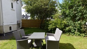 An outdoor patio with wicker furniture and a table, surrounded by a grassy lawn and a wooden fence. - Solferie Holiday Apartment Østerveien (Kristiansand)