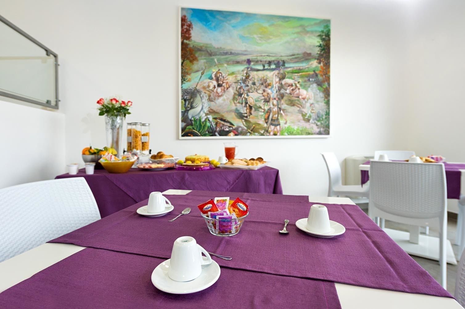 A dining area with a table set for breakfast, including cups, a bowl of fruit, and a painting on the wall.