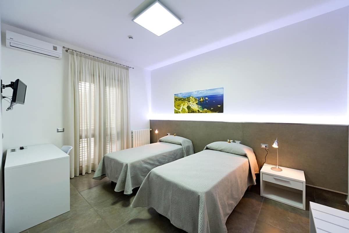 A hotel room with two beds, a desk, a chair, a radiator, a TV, and a painting of a coastal landscape.