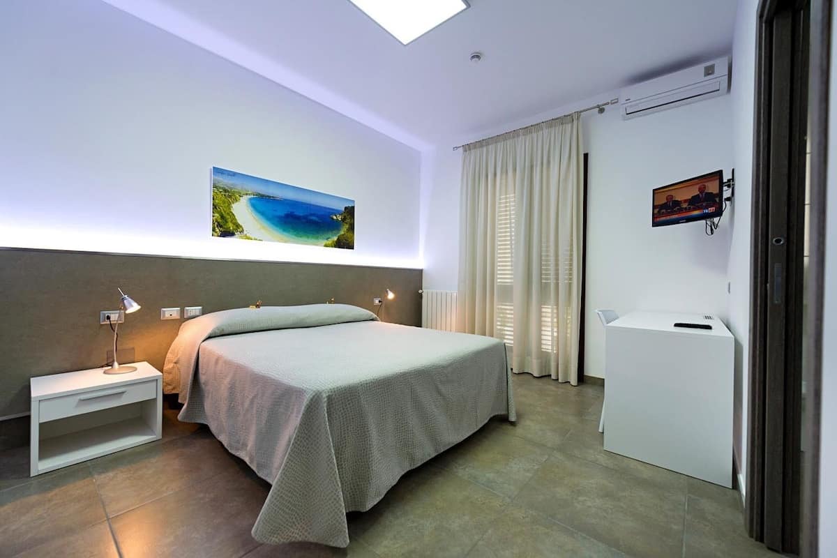 A modern hotel room with a bed, bedside table, lamp, and a painting of a beach.