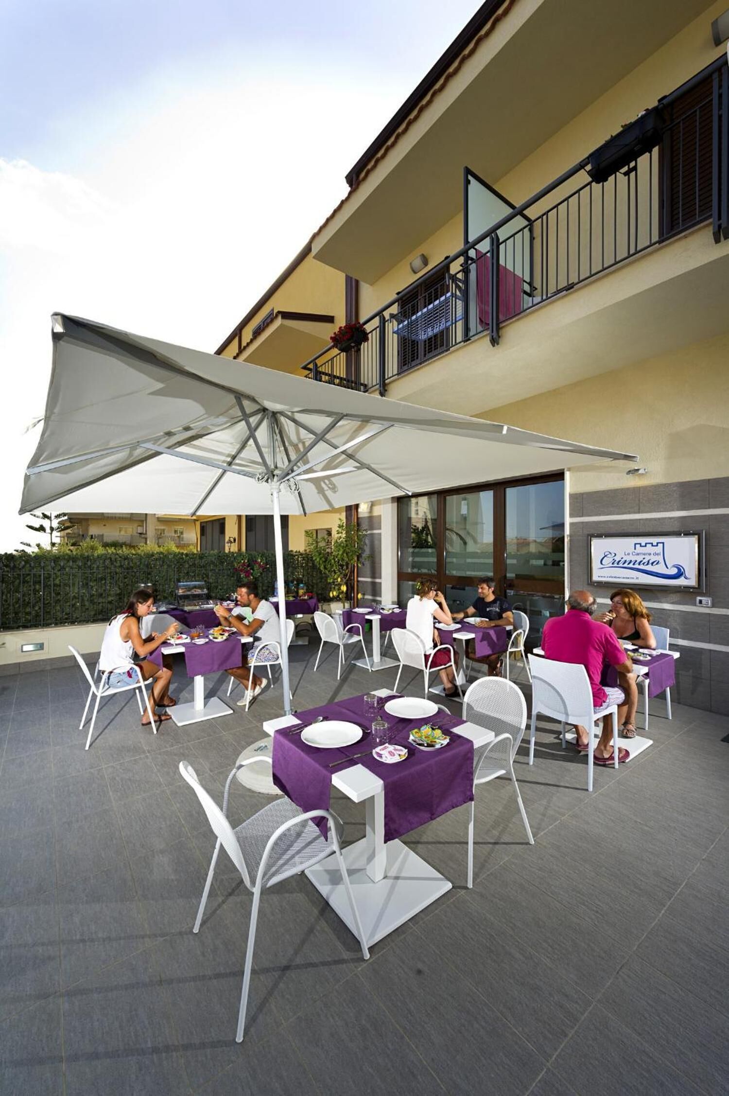 Een terras met tafels en stoelen, afgedekt door grote parasols, en een gebouw met een bord achtergrond.