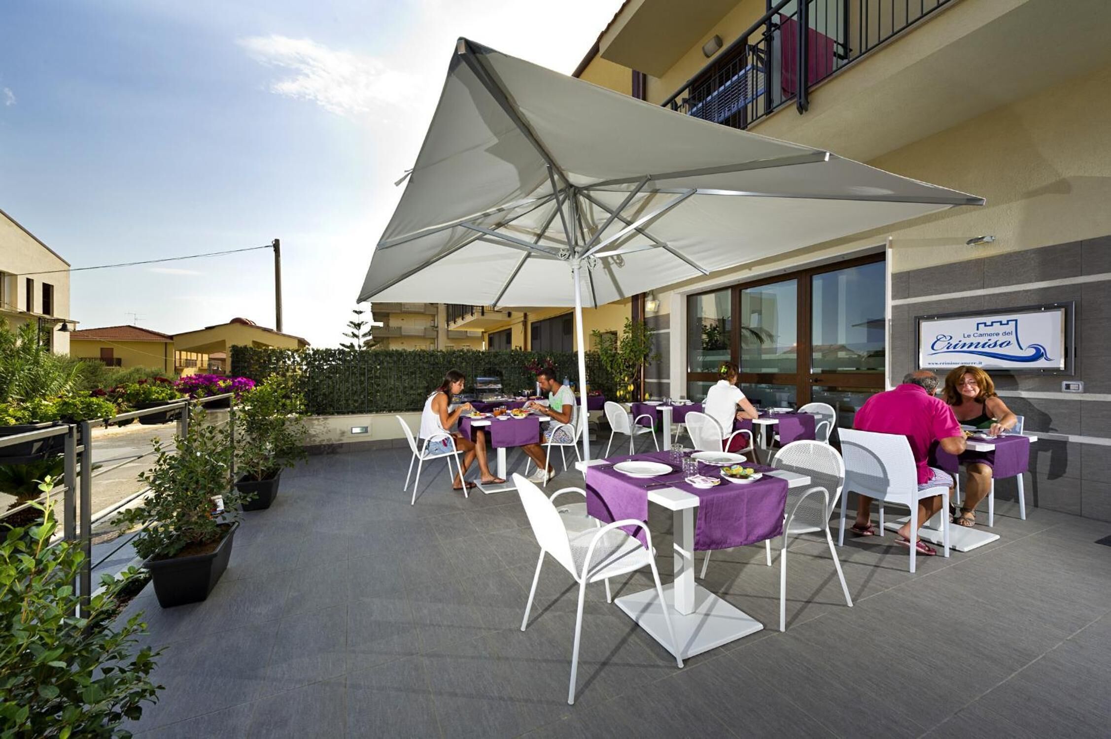 An outdoor dining area with tables and chairs, a large umbrella, and a sign for 'Le Camere del Cantiere'.