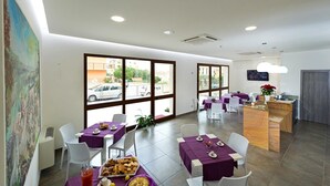 Un espace repas moderne avec des tables dressées pour les repas, de grandes fenêtres donnant sur la rue et un coin cuisine avec un comptoir en bois.