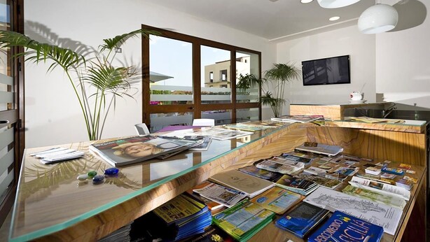 Un bureau moderne avec un bureau en verre, des livres et des magazines. On y trouve des plantes, une grande fenêtre et une vue sur les bâtiments environnants.