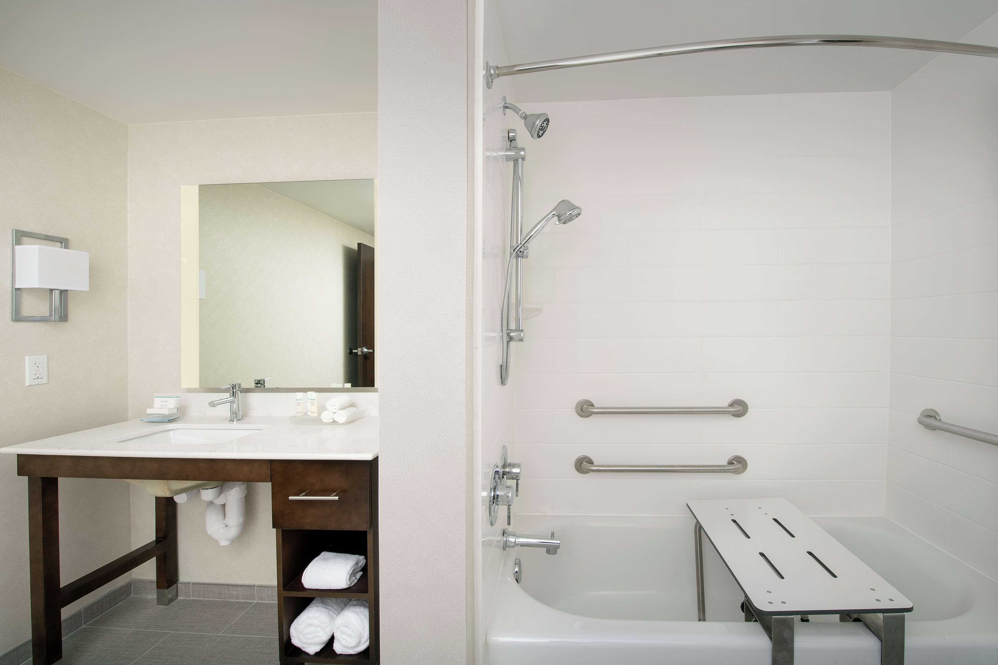 Een badkamer met een wastafel, spiegel, handdoekrek en een douche met handgrepen.