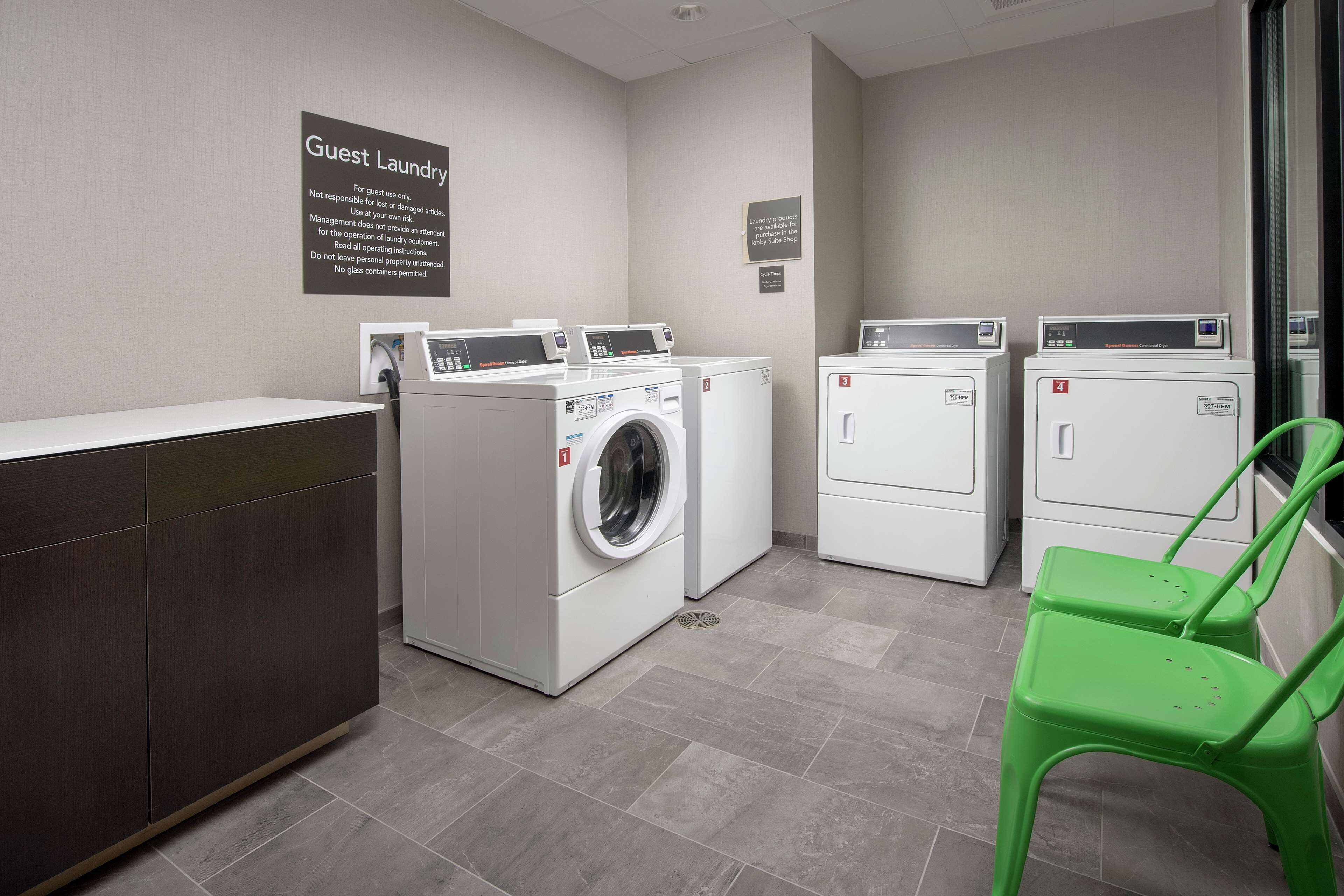 A guest laundry area with four washing machines, a green chair, and a sign indicating the use of laundry products in the lobby state shop.