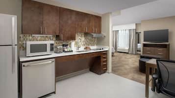 A compact kitchen with a microwave, refrigerator, and a small sink. There is a wooden cabinet above the counter and a television in the adjacent living area.