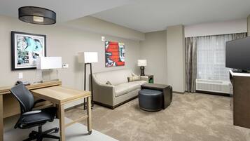 A hotel room with a desk, chair, sofa, ottoman, TV, and artwork on the walls.