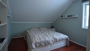 Ein Schlafzimmer mit schrägen Wänden, einem Bett mit gestreifter Bettwäsche, einem Nachttisch, einer kleinen Regalablage mit Büchern und einem Fenster.
