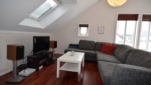 Ein modernes Wohnzimmer mit schrägverdeckter Decke, einem Flachbildfernseher, einem weißen Couchtisch und einem grauen Sofa mit roten Kissen.
