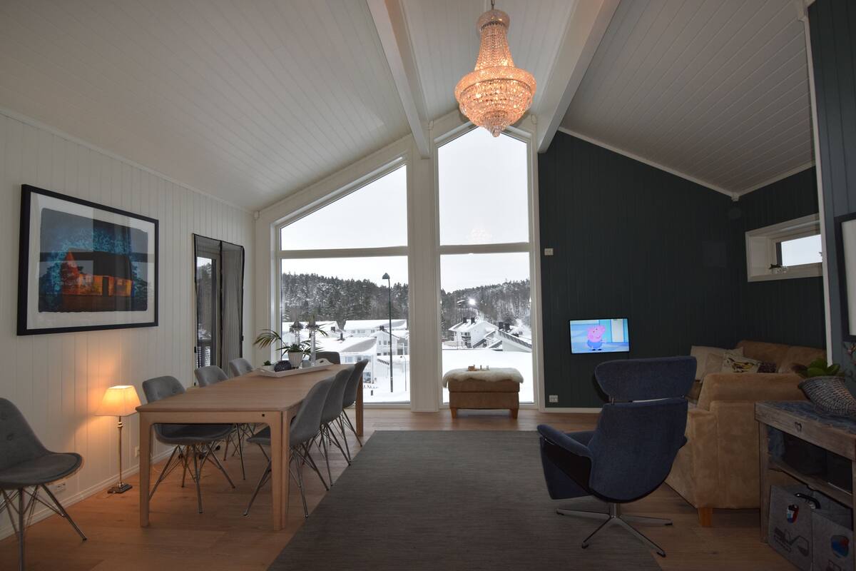 Una sala de estar moderna con una gran mesa de comedor, asientos cómodos, un televisor y vistas a un paisaje nevado a través de amplios ventanales.