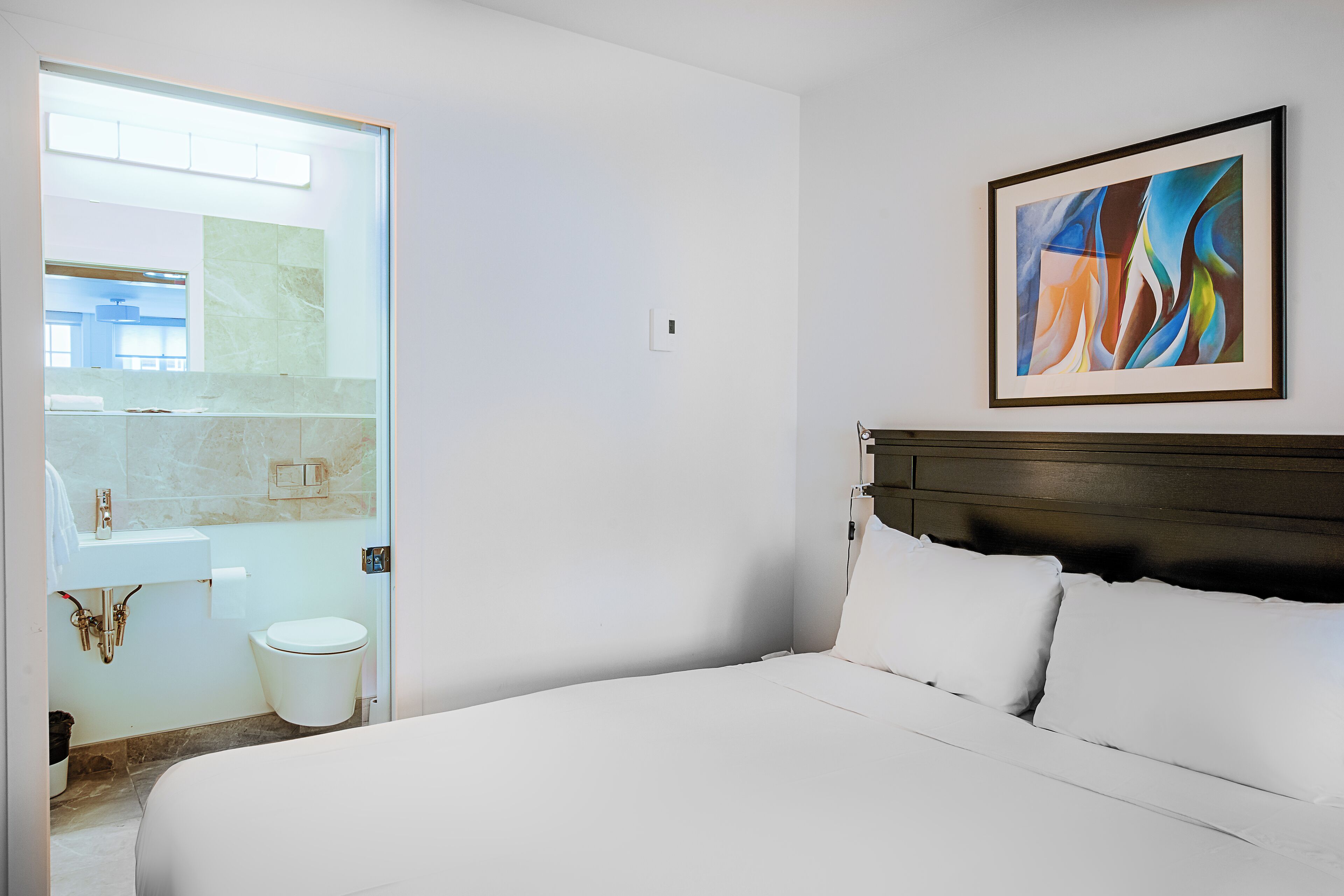A hotel room with a bed, a bathroom with a toilet and sink, and a painting on the wall.