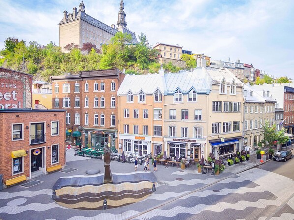 Un bâtiment historique doté d’une tour imposante, une place centrale avec une fontaine, et des immeubles commerciaux situés aux alentours.