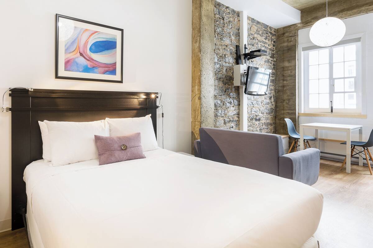 A hotel room with a large bed, a stone accent wall, a modern light fixture, and a window with a view.