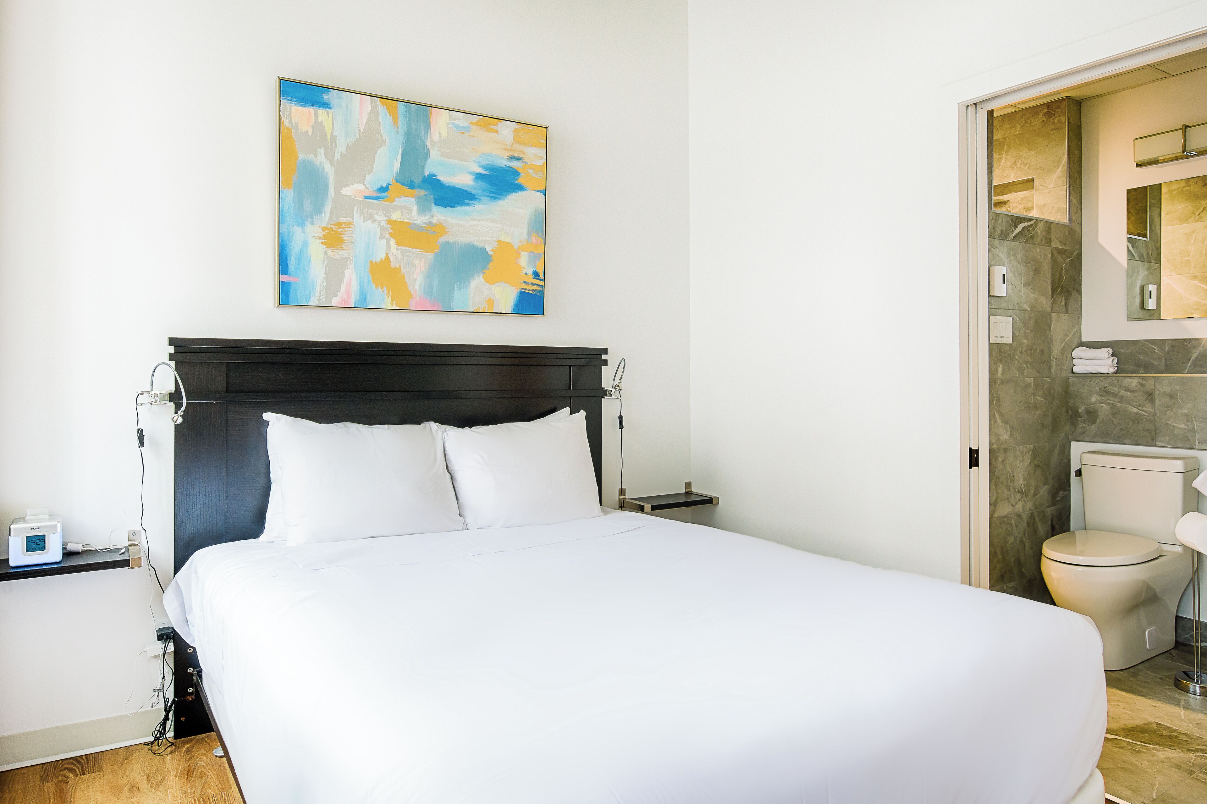A hotel room with a large bed, a nightstand, a wall-mounted abstract painting, a bathroom with a toilet and mirror, and a marble floor.