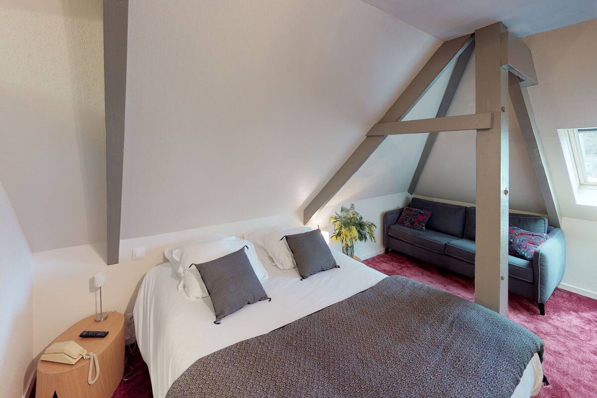 A bedroom with a sloped ceiling, a bed with white linens and patterned pillows, a grey sofa with a floral cushion, a small table with a lamp, and a window with a view of buildings.