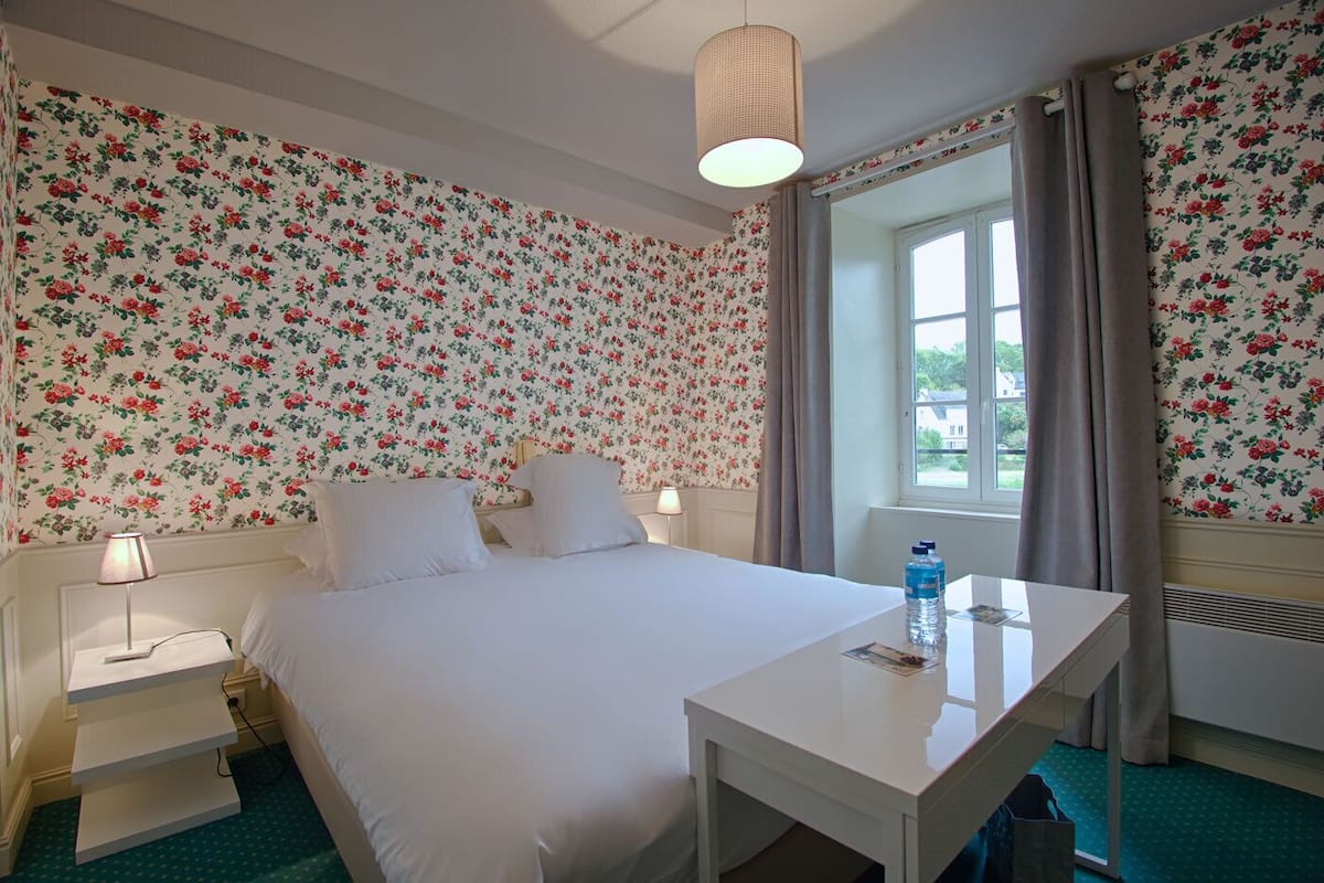 A hotel room with floral wallpaper, a bed, a desk, a lamp, and a window with curtains.