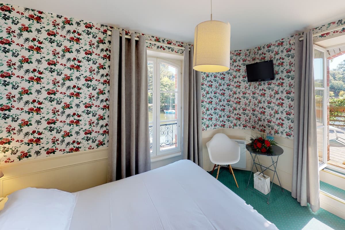 A hotel room with floral wallpaper, a bed, a desk with a chair, a television, and a view of the outdoors.
