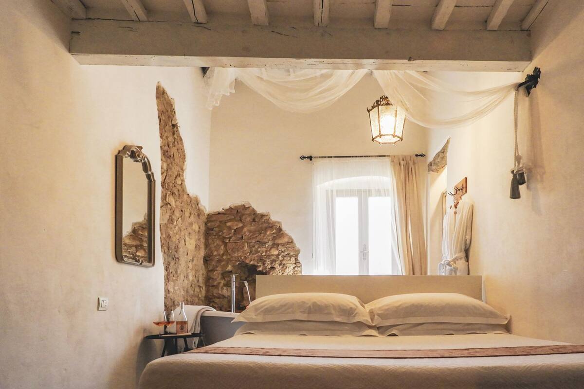A bedroom with a stone wall, a bed with white linens, a hanging light fixture, and a mirror.