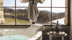 A bathroom with a bathtub, a window with a view of a vineyard, and a small table with a bottle and glasses. - Tenuta CorteDomina (Radda in Chianti)