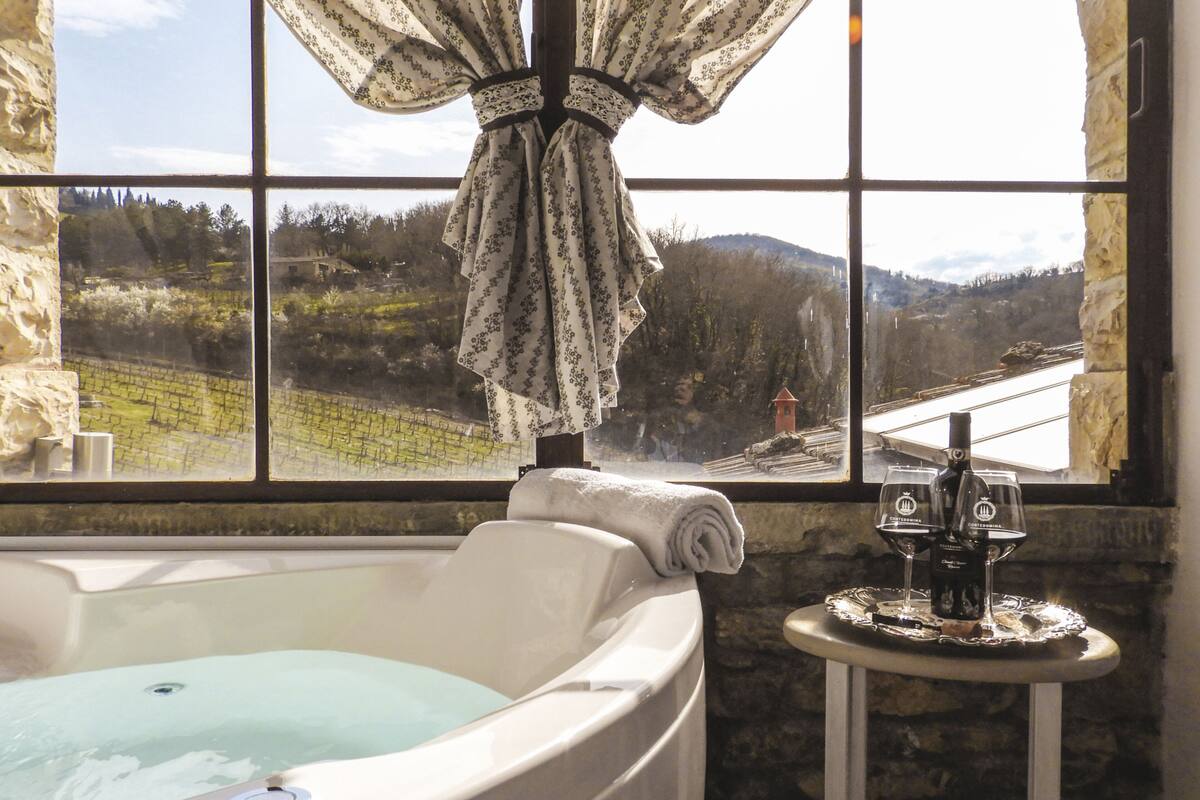 A bathroom with a bathtub, a window with a view of a vineyard, and a small table with a bottle and glasses.