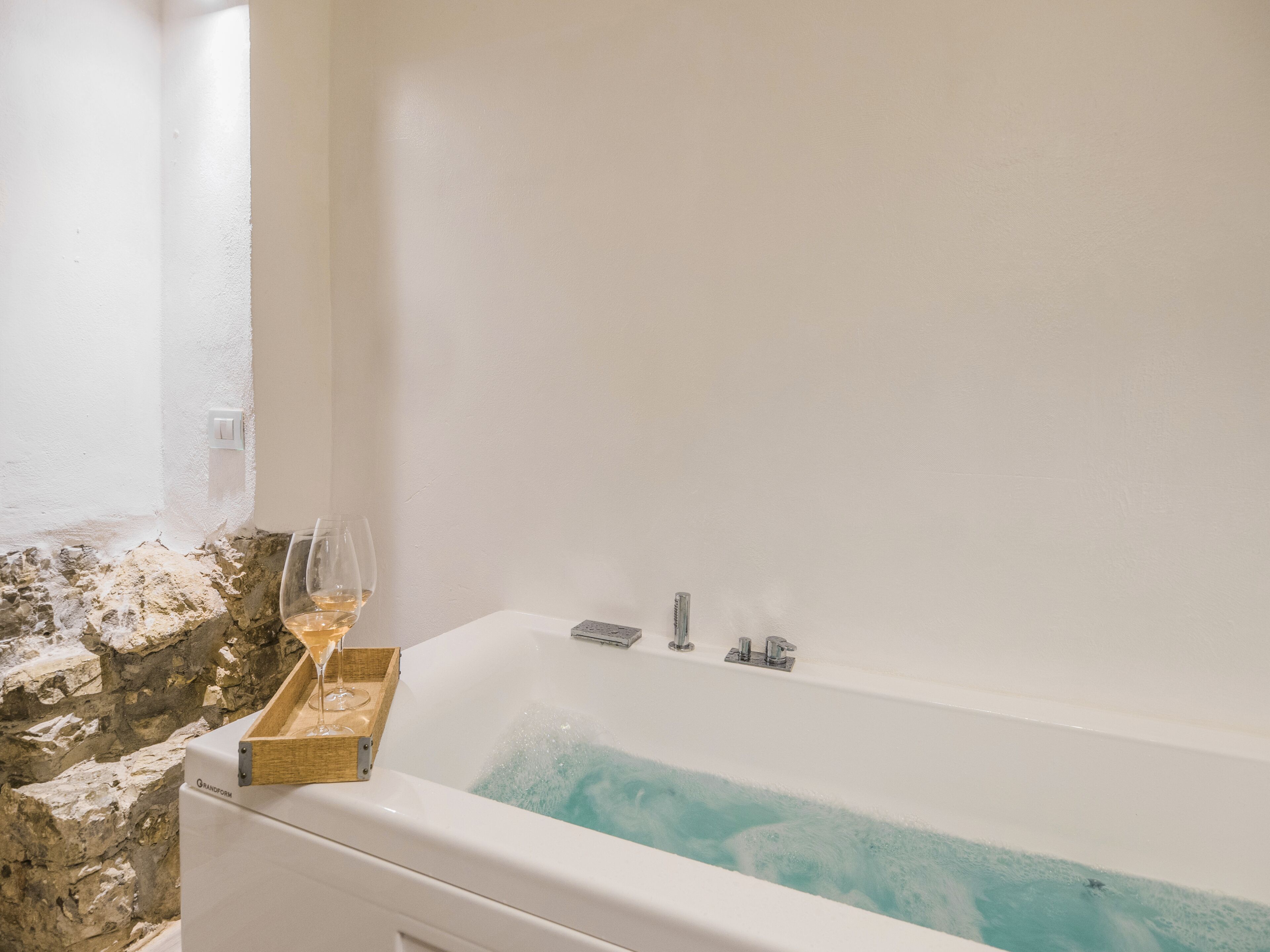 Une baignoire avec un plateau en bois sur lequel sont posés un verre et une bouteille, un mur en pierre et un mur blanc avec un interrupteur.