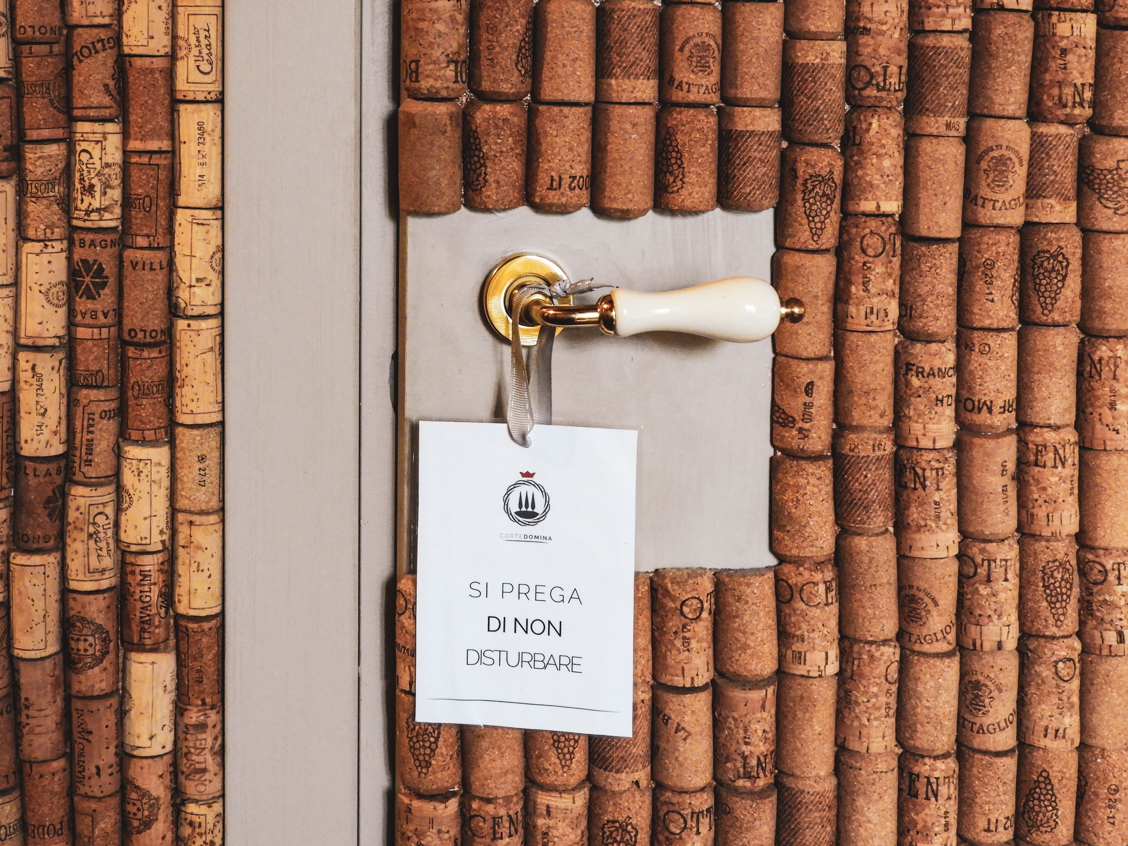 A door with a corkboard wall, a brass door handle, and a sign with the text "SI PREGA DI NON DISTURBARE".