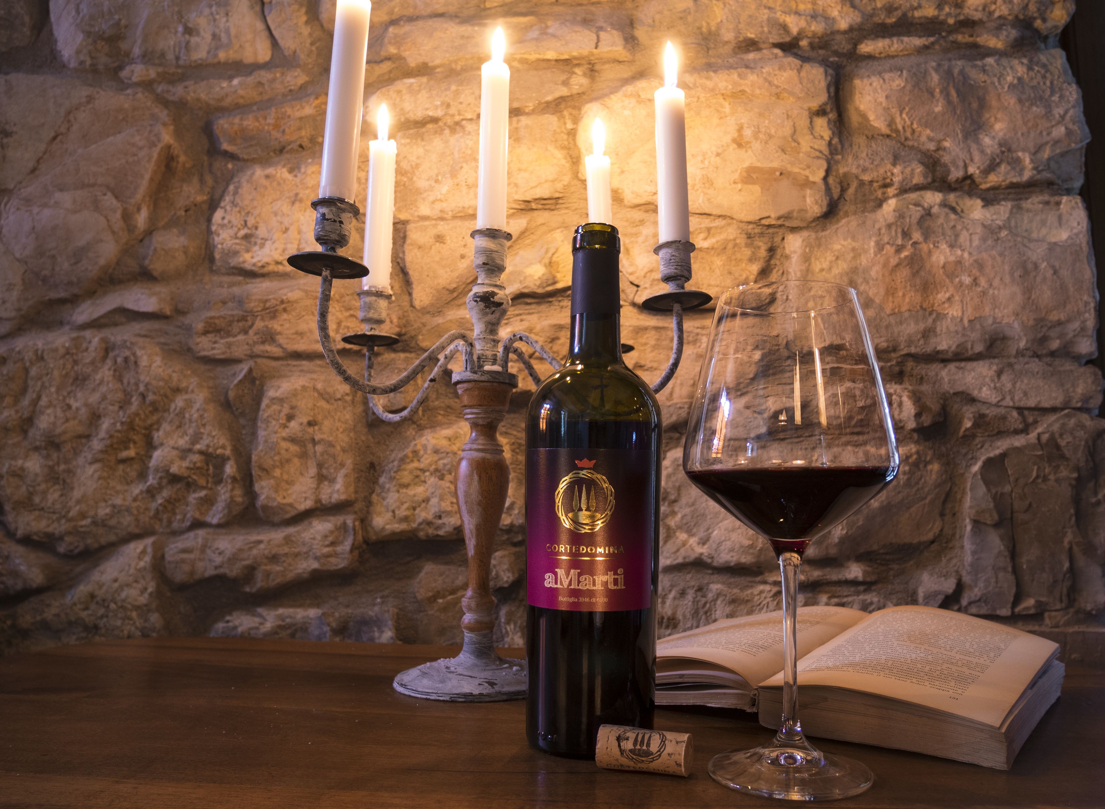A bottle of wine, a glass of red wine, a candle holder, and an open book on a wooden table.