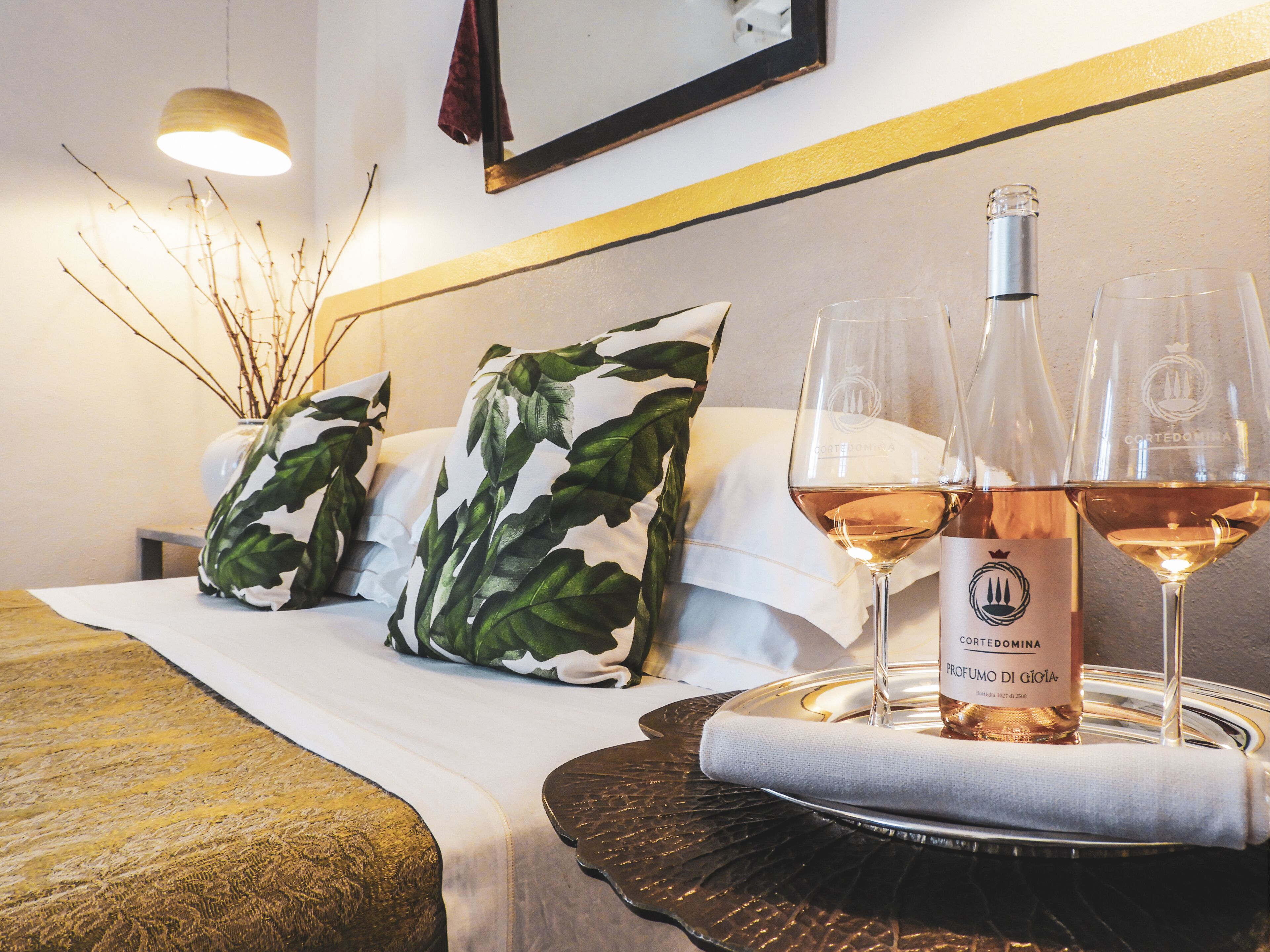 A neatly made bed with white linens, two pillows with a leaf pattern, a bottle of wine, and two glasses on a tray.