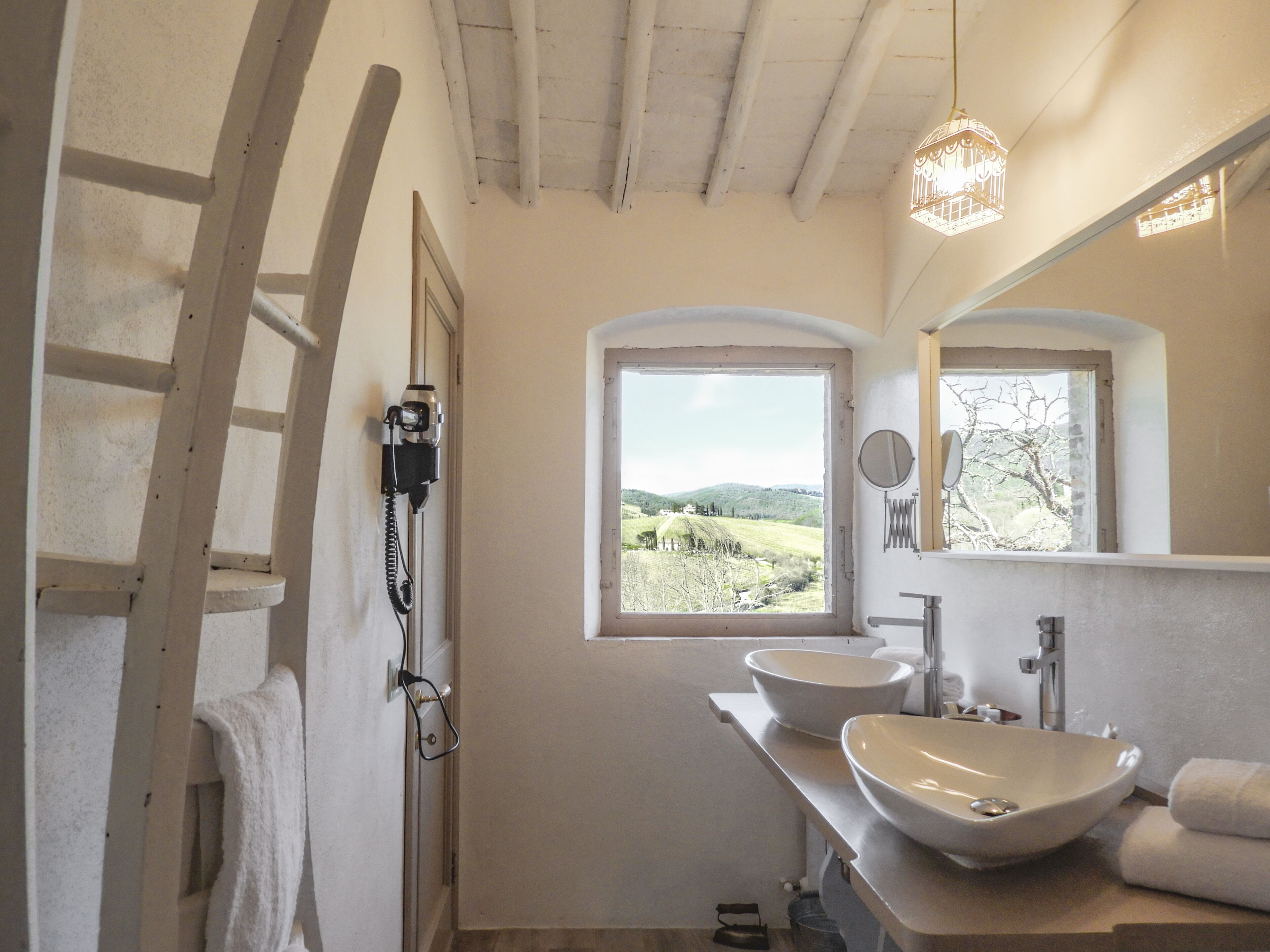 A bathroom with two large sinks, a ladder, a window with a view of hills, and a hanging light fixture.