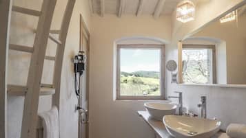 Une salle de bain avec deux grands lavabos, une échelle, une fenêtre donnant sur des collines et un luminaire suspendu.
