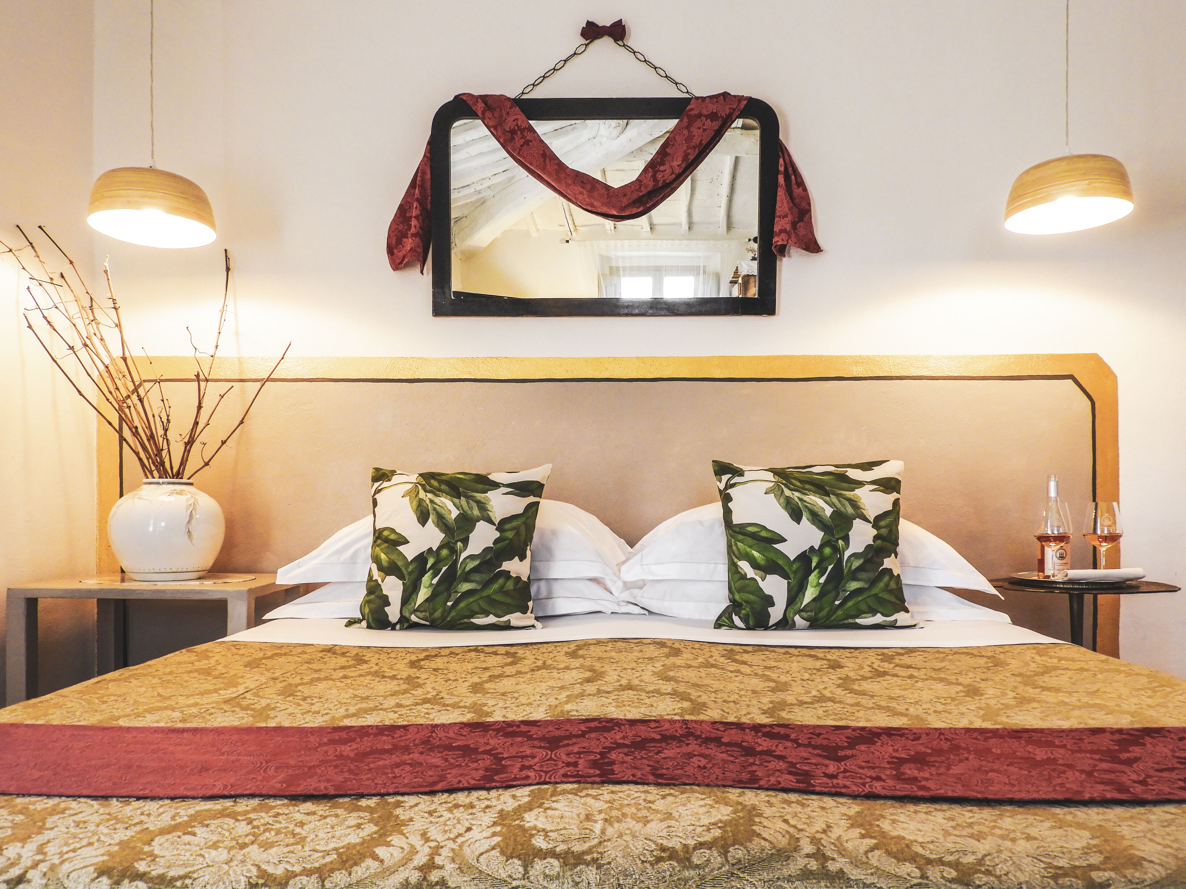 Una cama bien tendida con una colcha estampada, dos almohadas con diseño de hojas, un florero con ramas y un espejo con un paño colgante.