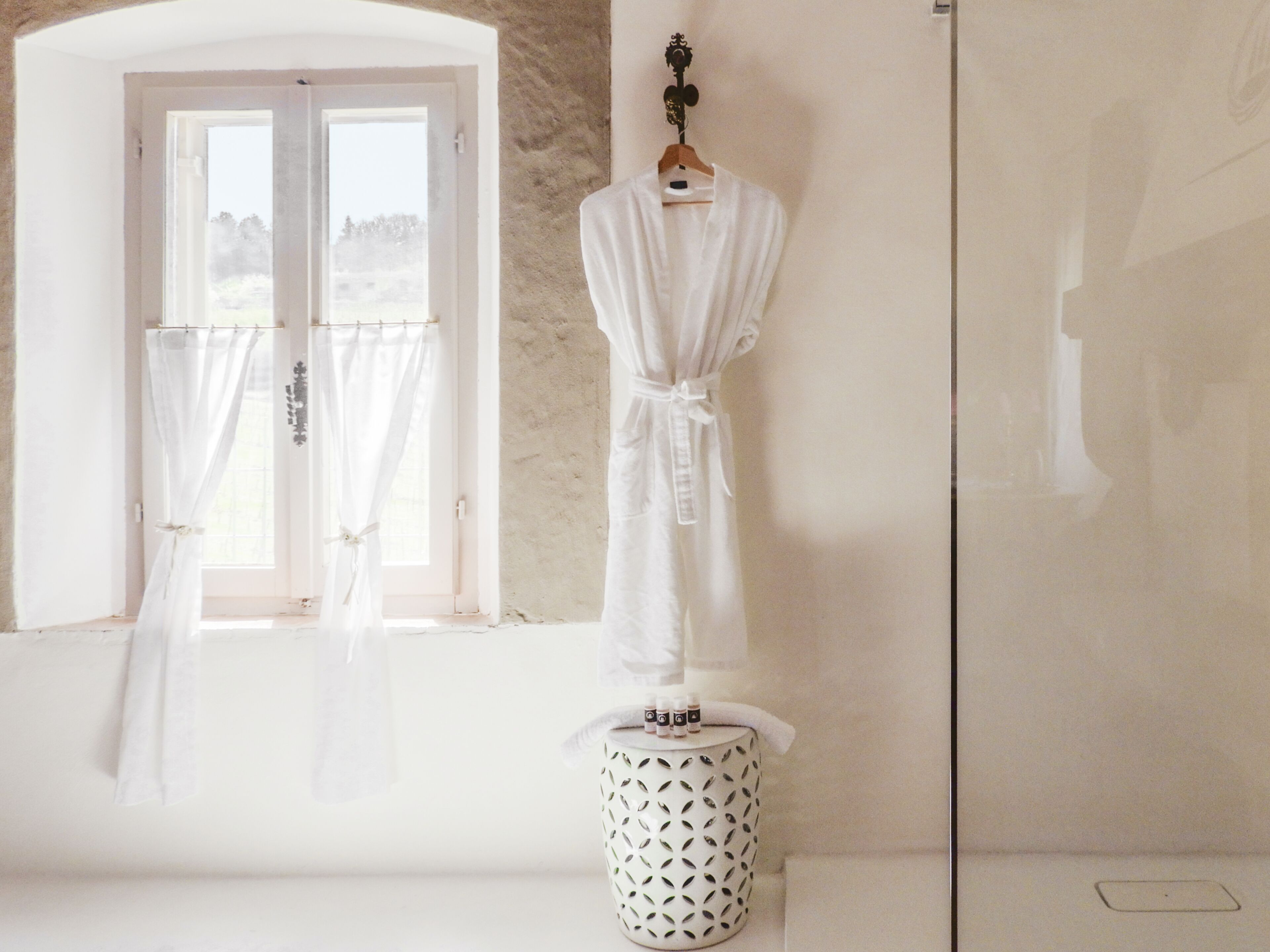 Une robe blanche suspendue à un cintre en bois, un tabouret blanc à motifs et une fenêtre avec des rideaux.