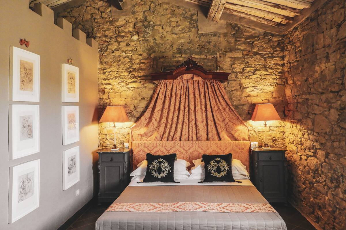 A bedroom with a stone wall, a wooden ceiling, a bed with a canopy, two bedside tables with lamps, and framed pictures on the wall.