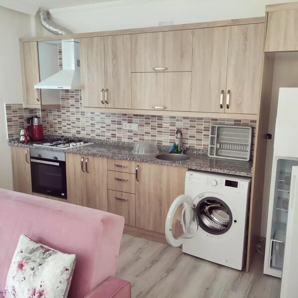 A compact kitchen with wooden cabinets, a built-in microwave, a washing machine, and a pink sofa with a floral pillow. - Villa Konak Yalcin (Ortaca)