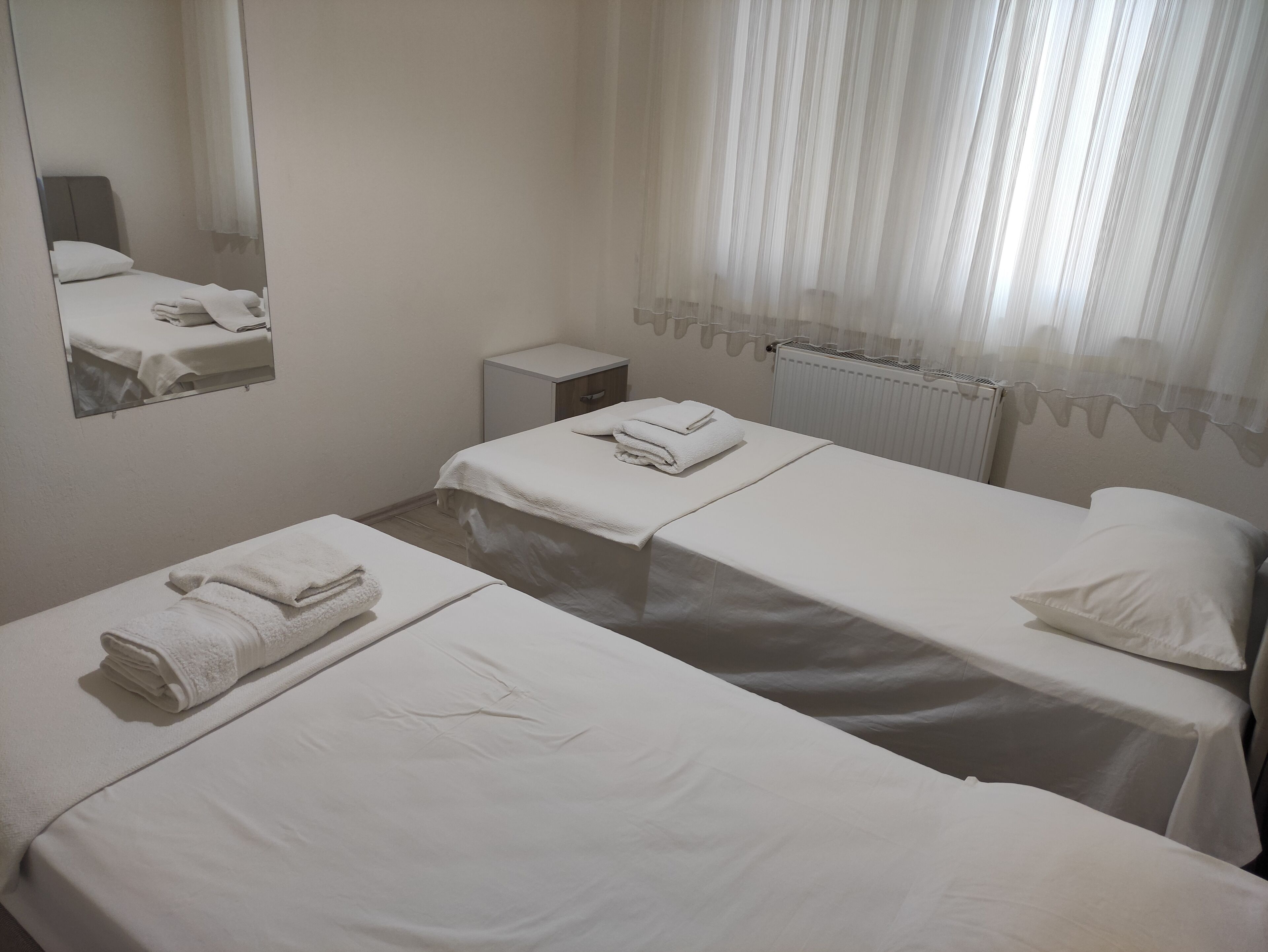 A hotel room with two beds, a small bedside table, a mirror, and a window with sheer curtains.