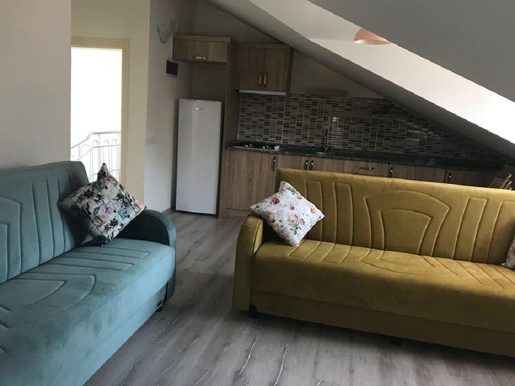 Un soggiorno con un divano e una poltrona, un angolo cottura con pensili e un frigorifero, e un soffitto a falde.