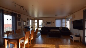 A spacious living area with a wooden dining table and chairs, a dark-colored sofa, and a flat-screen TV. - Solferie Holiday Home Marthas vei (Kristiansand)