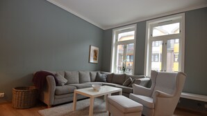A living room with a grey sofa, a white armchair, a small table, and a window with a view of buildings. - Stay.plus King Street (Kristiansand)